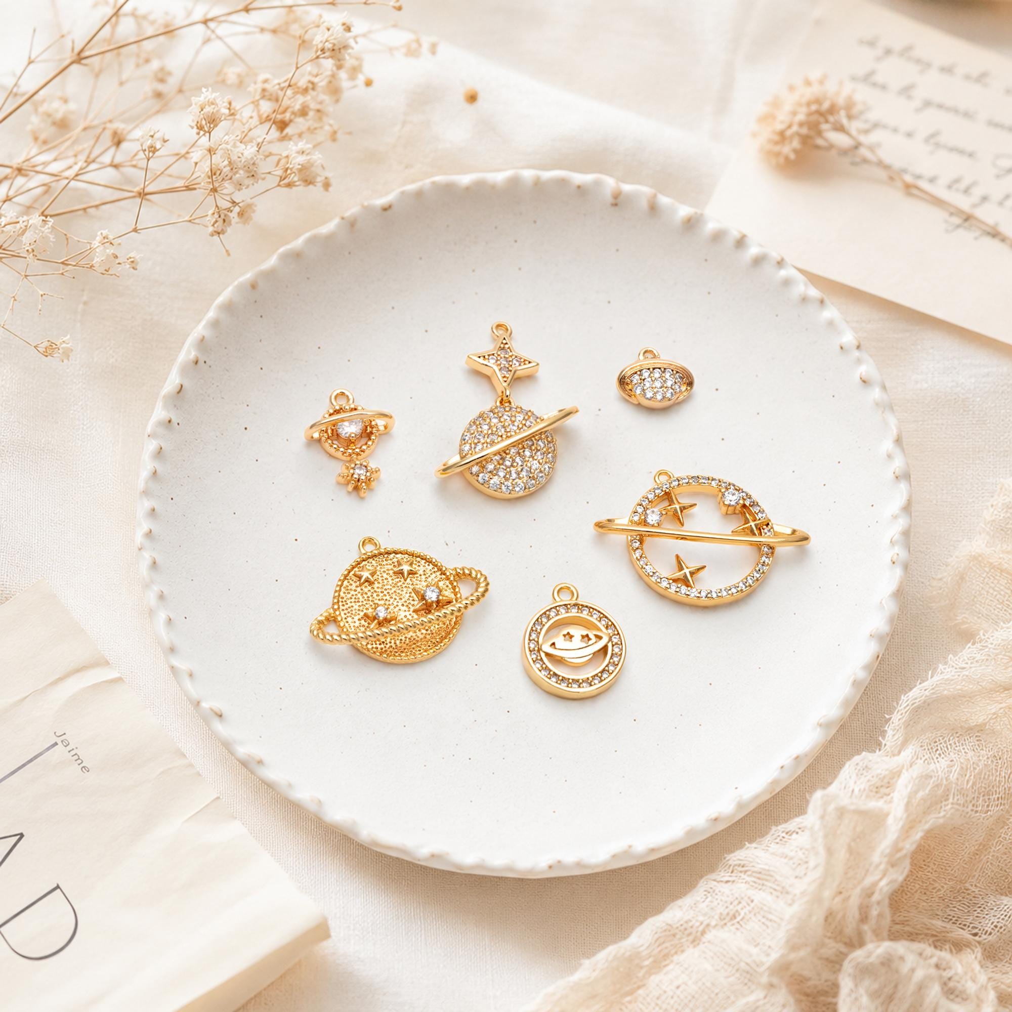 May include: A collection of six gold-toned metal charms shaped like planets and celestial bodies, featuring embedded clear crystals. The charms include various ringed planet designs, a star-topped planet, and a small oval shape, all with loops.